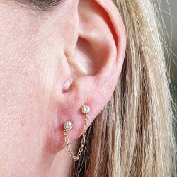 an ear lobe wearing two cz gems in gold from Anatometal with a chain hanging between the two